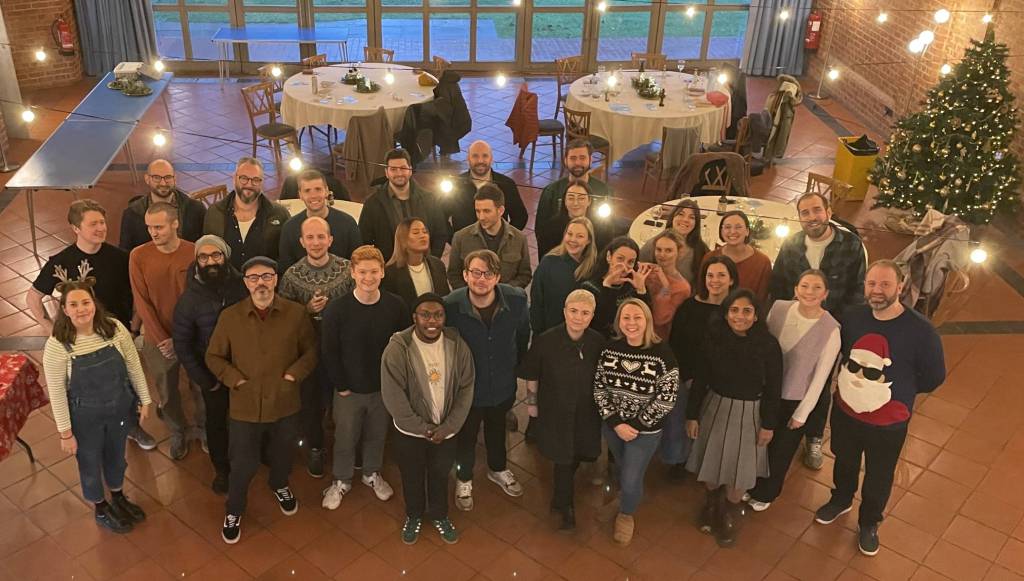 Photograph of the Substrakt team at their Christmas party. Taken from above, the team is all looking up at the camera. They are stood in a bunched group inside a festively decorated room. Everyone looks cheery.