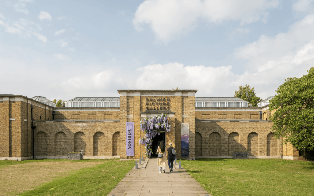 A photo of the Dulwich Picture Gallery building, taken in the daytime. Two people are walking down the path towards the building